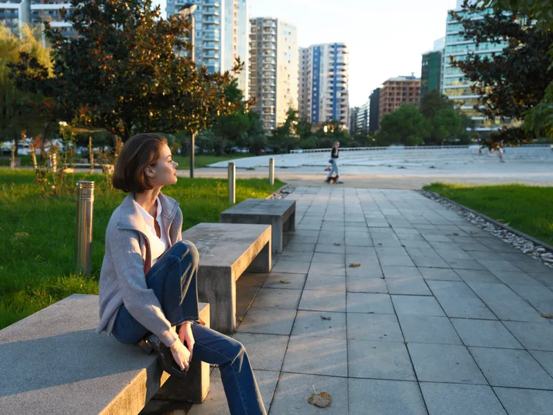 A contemplative woman sits on a concrete bench in a city park, bathed in warm sunlight. The calm, reflective mood is enhanced by distant skyline and trees, creating a peaceful urban moment.
