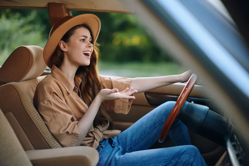 Happy young woman driving a car, wearing a stylish hat and casual outfit, expressing joy in a natural outdoor setting, emphasizing freedom and adventure.