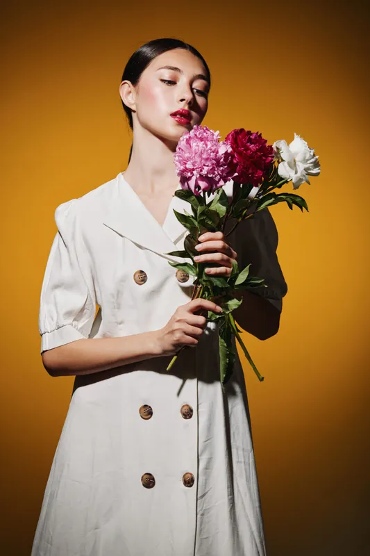 Young woman in white double-breasted dress holding pink, red, and white flowers against a vibrant orange background. Skin glows with natural makeup. Fashion and beauty concept.