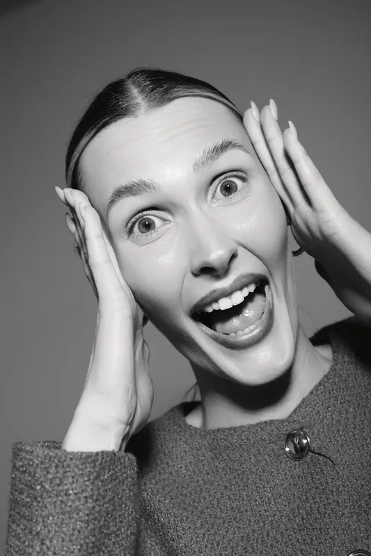 Excited Caucasian woman with light skin and pulled back hair expresses joy holding her face in both hands on grey background.