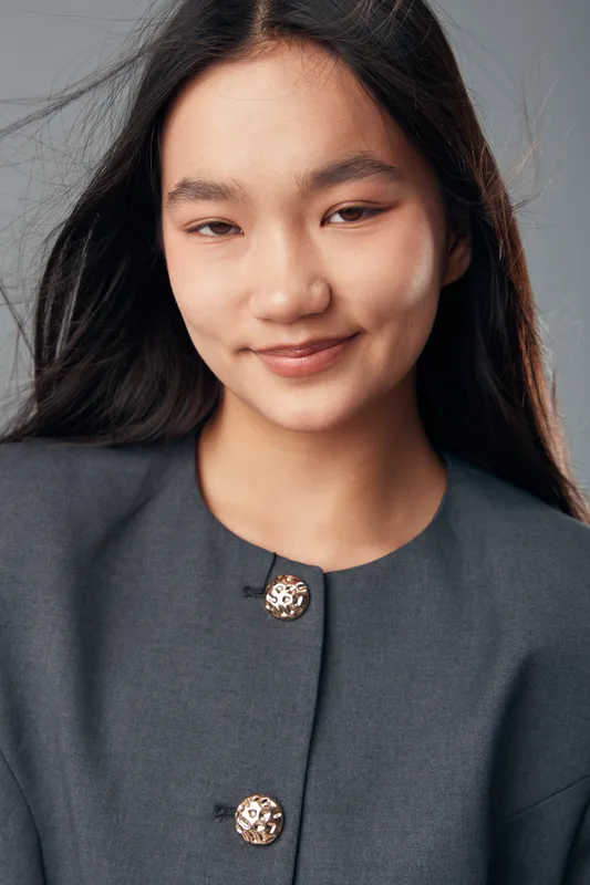 Portrait of smiling woman with long black hair wearing dark jacket with decorative buttons on grey background. Young adult female, beauty and confidence in studio lighting.