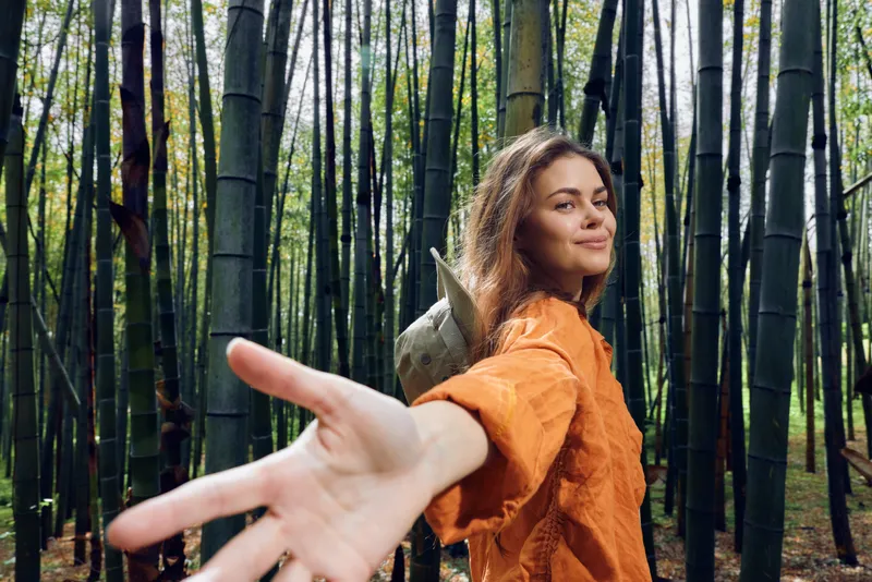 Woman in bamboo forest reaching hand toward camera, smiling with backpack and orange shirt. Hiking in nature among tall green trees, peaceful outdoor adventure and exploration.