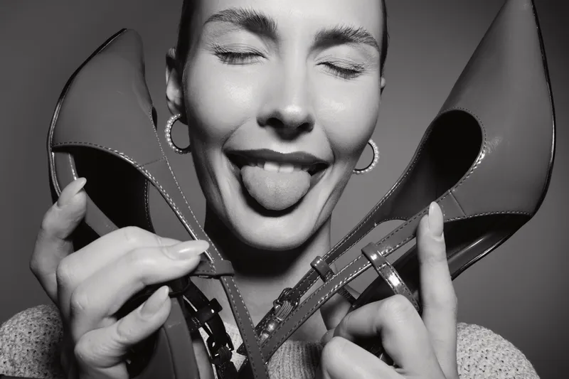 Playful white woman with light skin holds red shoes near face, sticks out tongue, smiling with closed eyes in black and white portrait. Concept of fun and fashion.