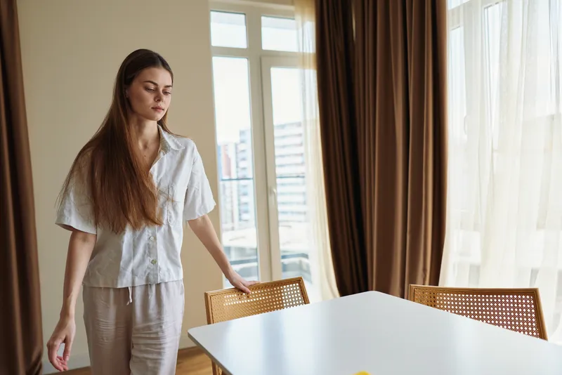 Woman at home by table and window with chair and curtains, natural light fills modern interior as a casually dressed young woman stands calmly next to dining table.