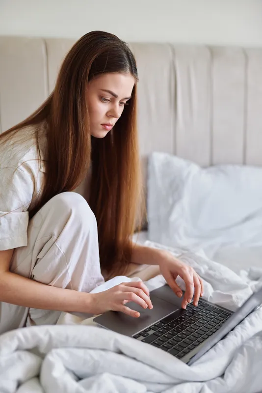 Woman with laptop on bed in a softly lit bedroom, wearing pajamas and working with focused concentration, typing on keyboard and handling remote tasks in a cozy home setting.