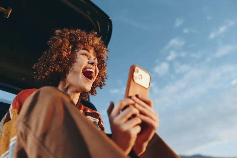 Cheerful woman lifestyle outdoors, laughing while taking a selfie with a smartphone, sunlit moment, curly hair in the breeze, blue sky background, warm natural colors