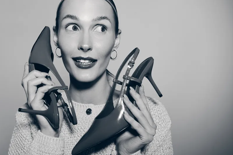 Young white woman with fair skin holds red high heel shoes near her face, smiling and looking aside. Concept of fashion, shopping, style, and footwear choice.