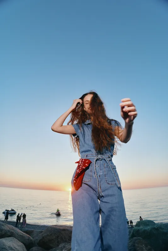lifestyle woman in modern boho-western denim outfit with long hair enjoying sunset by the sea, film color tones capturing casual street style vibes