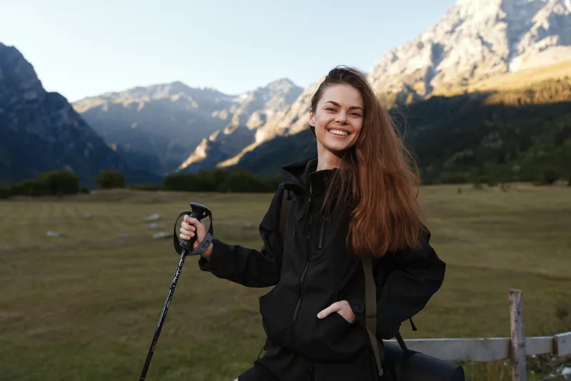 A smiling young traveler stands outdoors in a mountainous landscape, wearing a dark jacket and holding a trekking pole. The scene conveys confidence, adventure, and optimism, capturing a vibrant