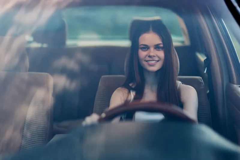 Happy young woman smiling while driving a car, showcasing joy and freedom, with an inviting interior backdrop and warm color tones.