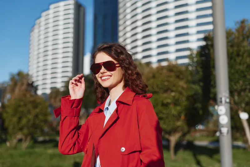 Woman in a red trench coat with sunglasses smiles in a sunny park, modern high rise buildings visible behind, creating an urban chic mood.
