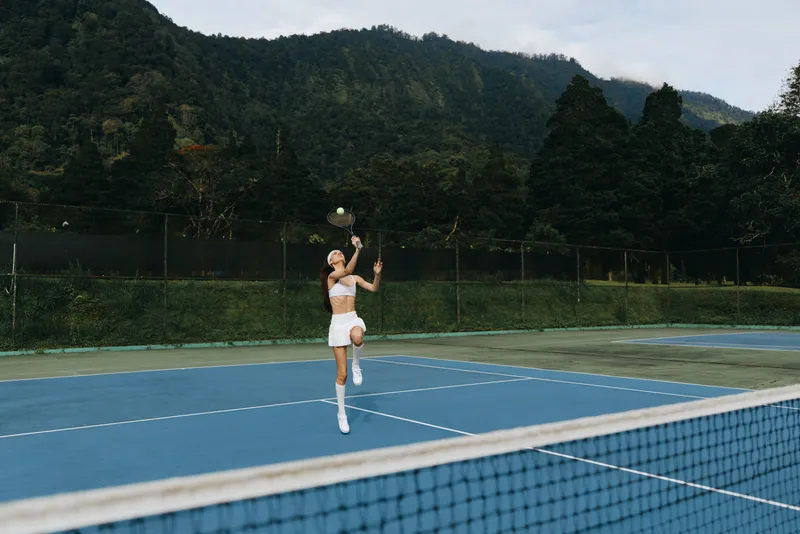 Young Asian woman playing tennis on a sunny day, demonstrating joy and energy on a vibrant blue tennis court surrounded by nature