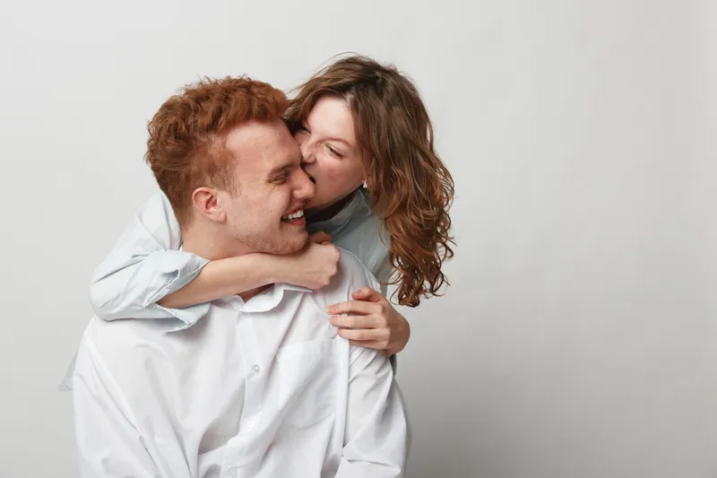 Happy young couple in white shirts share affectionate moment, woman hugging and kissing man on cheek. Studio portrait on light background. Concept of love, intimacy, and relationship happiness.