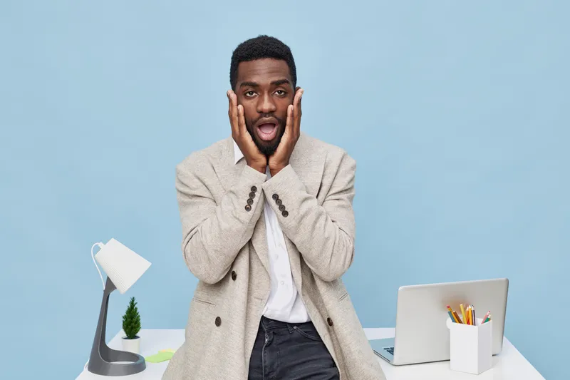 Joyful young man with surprised expression, hands on face, wearing beige patterned sweater, standing at modern workspace with laptop and desk lamp, on blue background, studio portrait, human emotion c