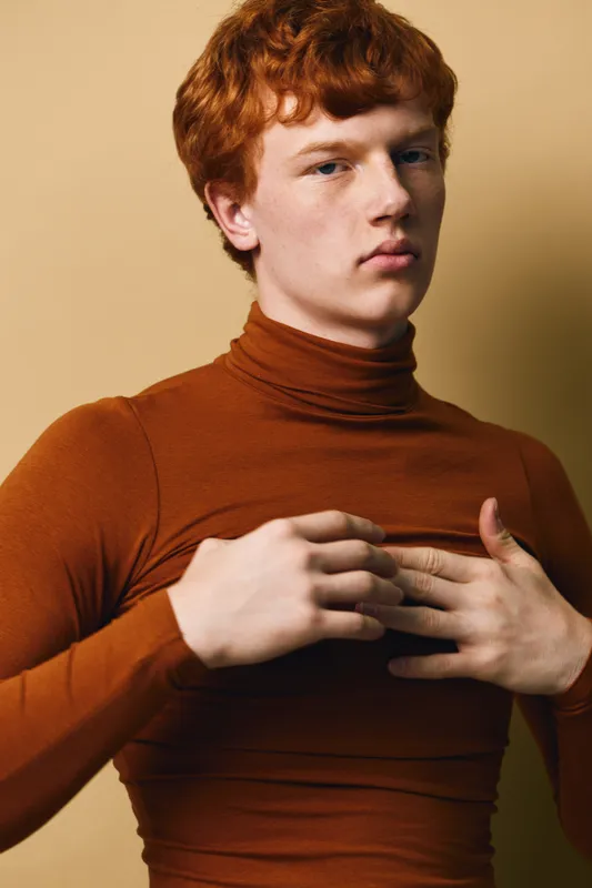 Young man with red hair wears brown turtleneck sweater and adjusts clothing while looking at camera in studio with beige background. Portrait of caucasian male model.