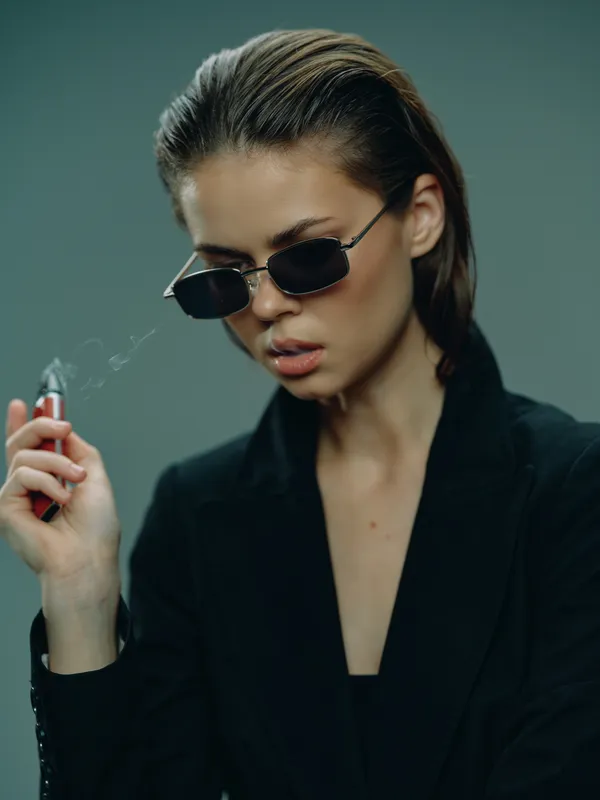 Stylish woman wearing sunglasses smoking electronic cigarette with serious expression in dark outfit and moody background