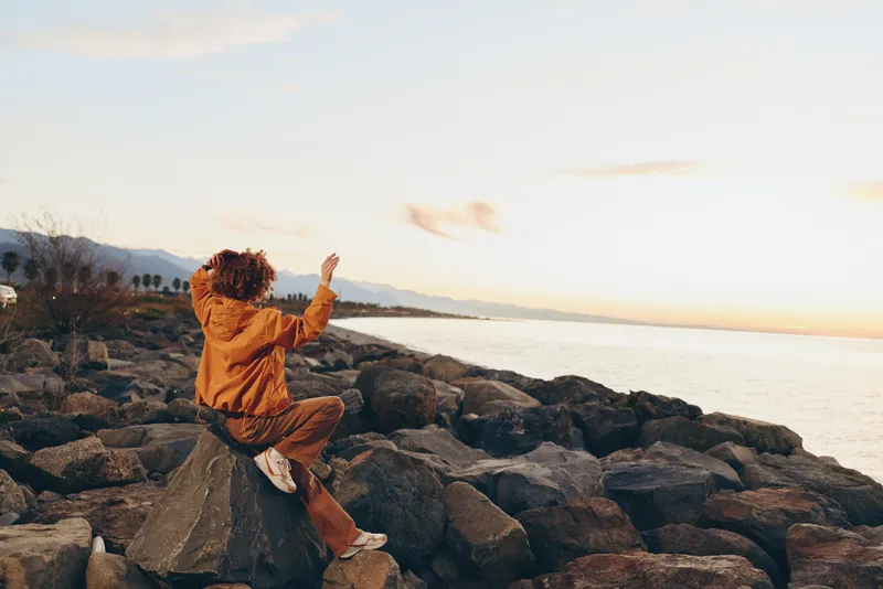 Woman lifestyle portrait on rocky beach at seaside sunset, arms raised, gazing toward ocean horizon, along the coast, travel and vacation concept in warm golden light