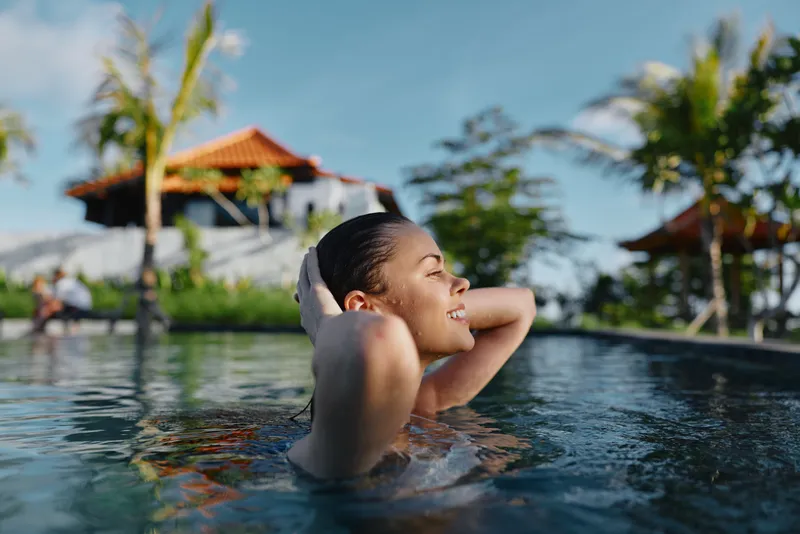 Cheerful woman enjoys poolside relaxation outdoors, sunlight on her face, serene water, tropical vibe, casual mood, warm breeze, vibrant colors, calm leisure moment