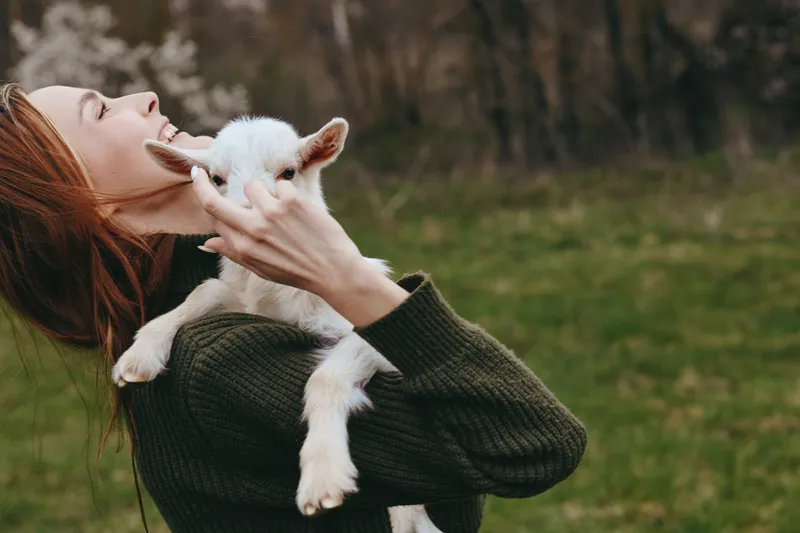 Young woman with long red hair joyfully holds a white baby goat close to her face in a green field during a casual outdoor lifestyle moment.