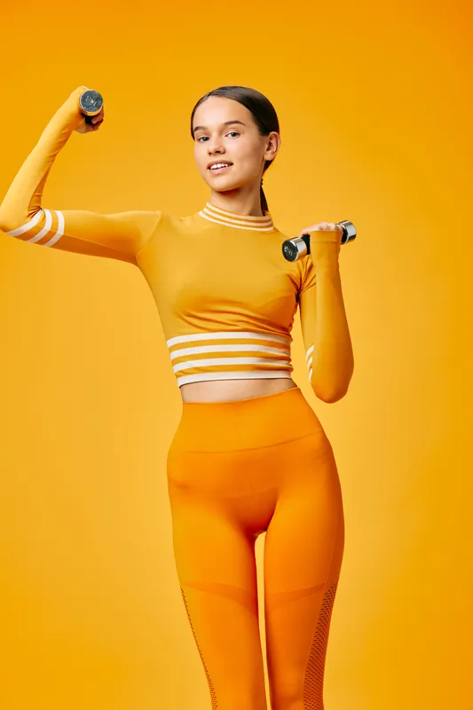 Fitness woman in bright orange outfit lifts dumbbells in a vivid orange studio, showcasing athletic strength, balance, and focused expression during a dynamic workout.