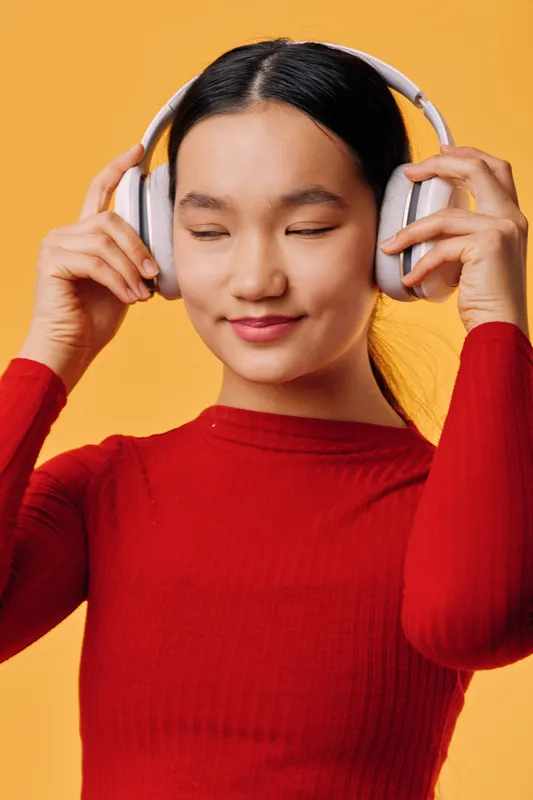 Young woman with headphones smiling and enjoying music with closed eyes. Female in red sweater listens to sound content on orange background with relaxation and calmness.