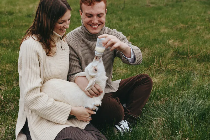 couple outdoors goat kid feeding bottle meadow grass joyful moment sunlit countryside warm sweaters candid scene natural colors relaxed lifestyle affectionate interaction wholesome family vibe