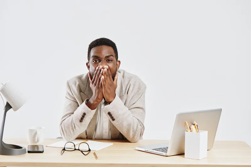 Honest african american man in beige blazer sitting at office desk covering mouth with hands looking surprised or shocked in front of laptop and documents workplace interior concept