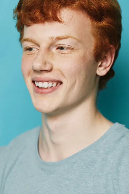 Young man with red hair smiling and looking away in closeup portrait against blue background. Casual style with light grey shirt and natural expression for positive mood.