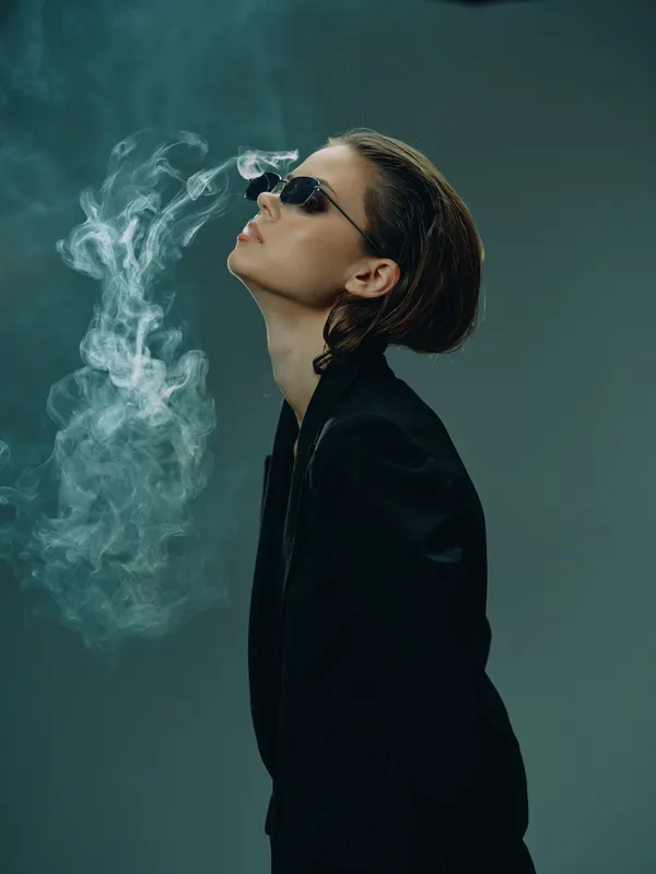 Stylish woman in black jacket wearing sunglasses exhales smoke against a dark background, creating a moody artistic atmosphere.