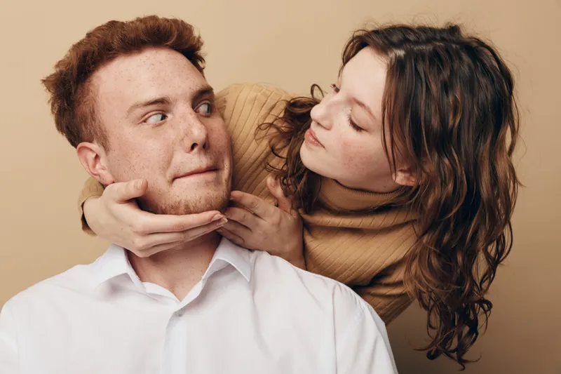 Young woman in brown sweater playfully holds young man in white shirt by the chin, both with expressive faces on beige background. Concept of fun, relationship, and playful interaction.