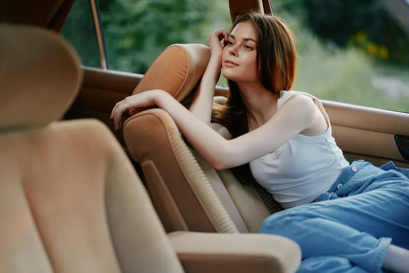 Beautiful young woman relaxing in a car interior, wearing a white tank top and denim jeans, expressing calm emotions in a nature-filled background.