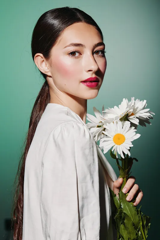 Young woman with glossy dark hair and bright makeup holds white daisies in hand against blue background. Beauty, fashion, flower bouquet, natural style, and calm expression concept.