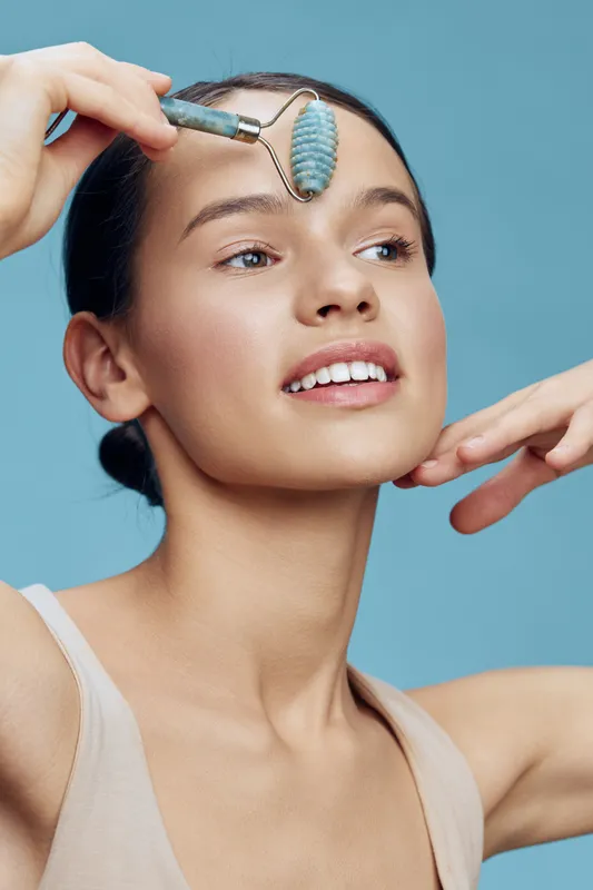 Young woman using a jade roller for skincare, showcasing a healthy glow and relaxation against a soft blue background Skin care, beauty, wellness, and self care concept