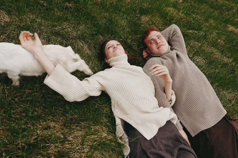 A young woman and man lie on grass wearing knitted sweaters while a white baby goat sniffs the woman's hand in a relaxed outdoor lifestyle scene.