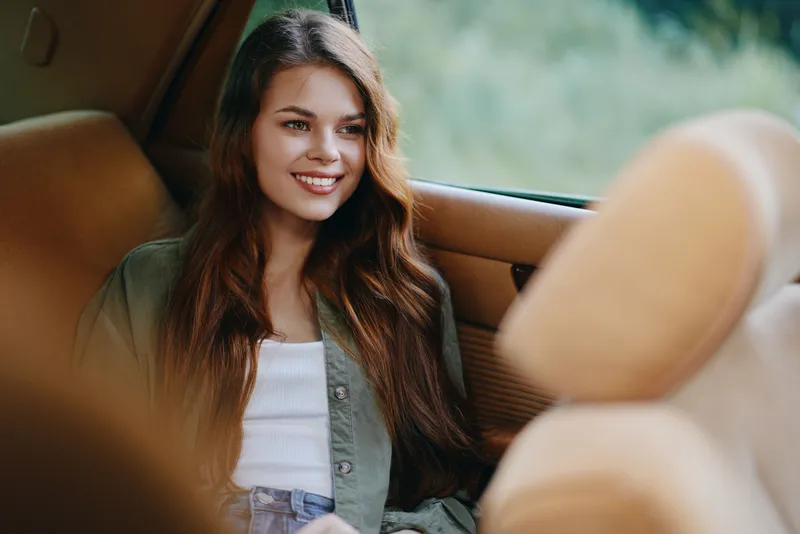 Young woman with long hair sitting in a car, smiling cheerfully, wearing casual green jacket and white top in a natural outdoor setting.