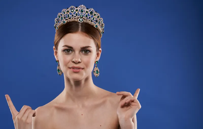 Женщина с обнаженными плечами и диадемой на голове жестикулирует руками на синем фоне