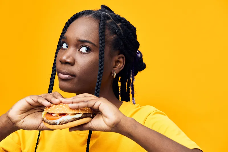 Young Black woman with braids holds a delicious hamburger, expressing curiosity and joy against a vibrant yellow background Fast food concept