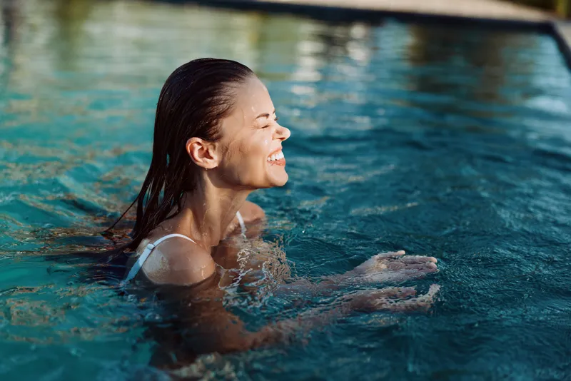 A smiling woman swims in a bright pool, enjoying a refreshing moment outdoors. The scene conveys relaxation, wellness, and carefree leisure in a sunny poolside setting.