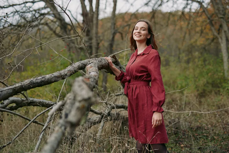 Smiling woman in a simple red dress stands beside a fallen tree in a woodland, conveying natural authenticity and quiet confidence through candid pose, texture, and warm, soft light.