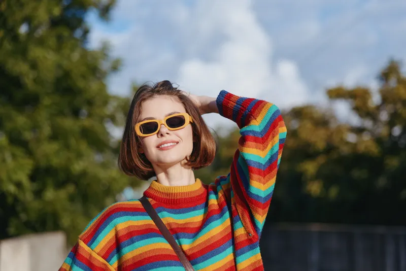 Woman in rainbow sweater and yellow sunglasses smiling outdoors, young adult with short bob haircut, casual crossbody bag, posing hand through hair in sunny park for lifestyle fashion travel.