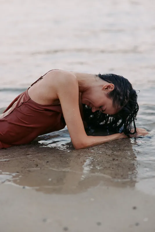 Young woman with wet hair resting on sandy beach, wearing brown dress, relaxed and peaceful, natural light, calm water in background.