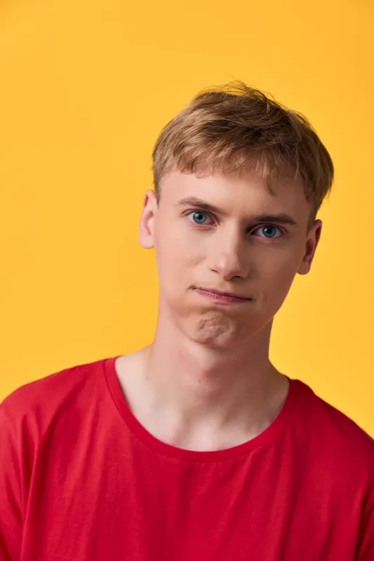 Puzzled, confused expression on a young man wearing a red shirt against a bright yellow background, conveying uncertainty and curiosity in a simple studio shot.