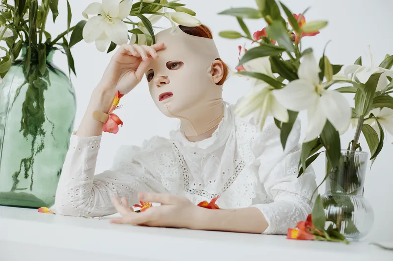 A contemplative model with a mask poses among white lilies, showcasing beauty and vulnerability in a serene, artistic composition ideal for wellness concepts