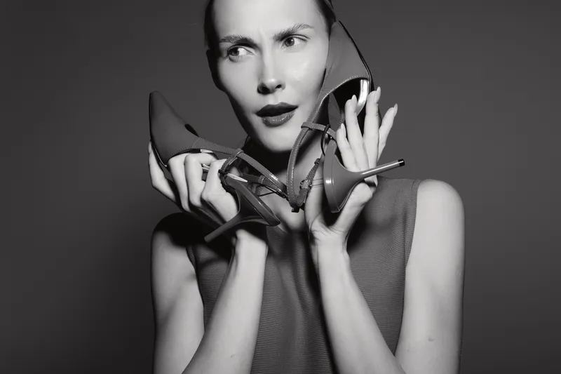 Young white woman with pale skin holding two high heel shoes near face, looking puzzled in black and white portrait.