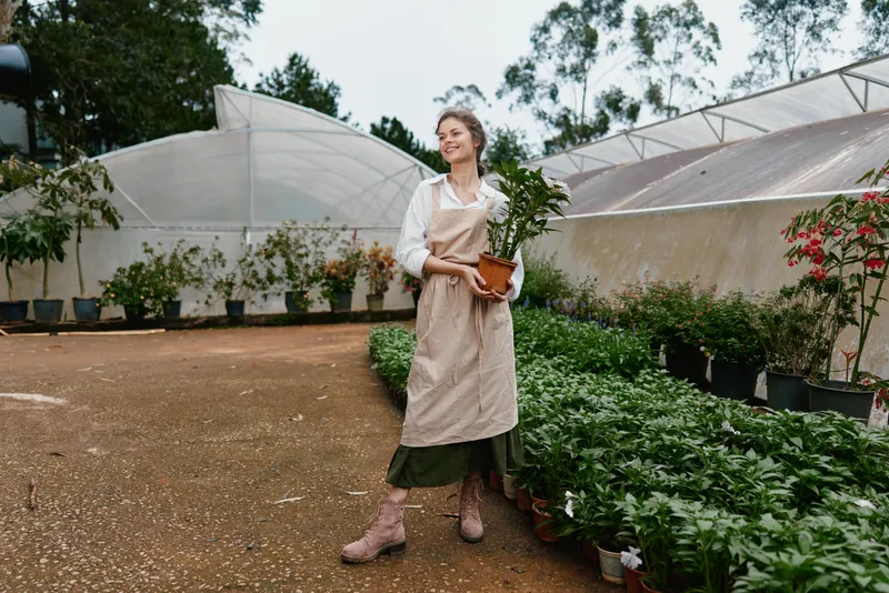 Young woman in gardening apron holding a plant in a greenhouse, showcasing a connection with nature and healthy living