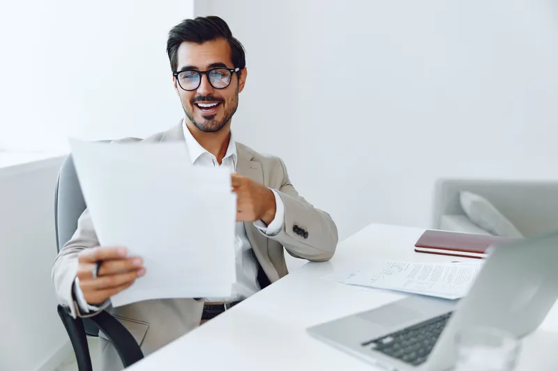 Улыбающийся бизнесмен в костюме держит документ и работает за ноутбуком в офисе