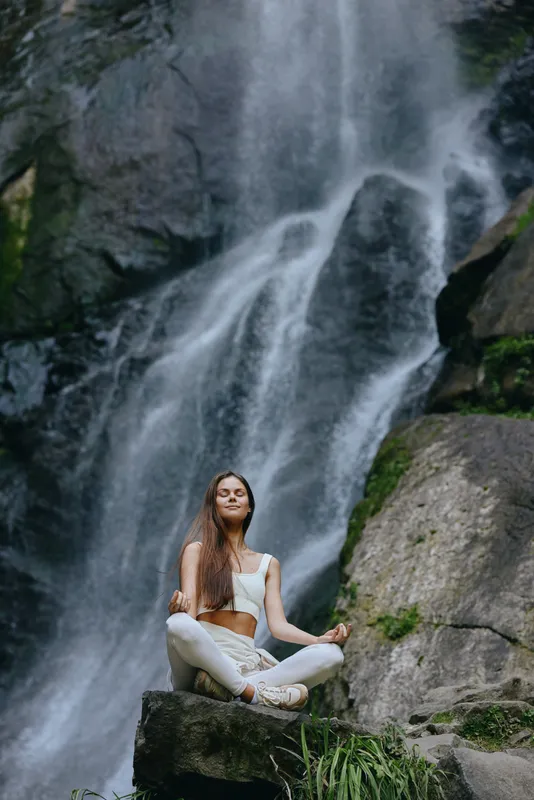 Young woman meditating outdoors near a natural waterfall, wearing white clothes, calm and peaceful expression, surrounded by rocks and greenery in serene nature setting.