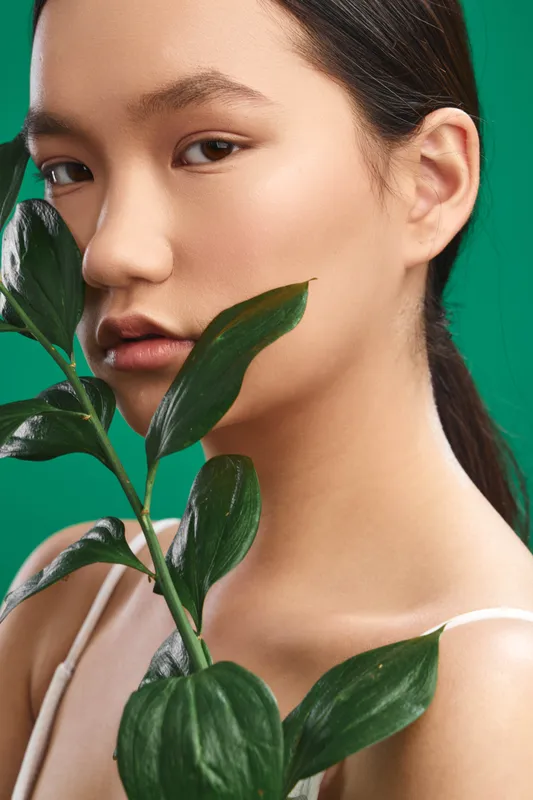Asian woman is captured in a closeup portrait with lush plant leaves and a vivid green background. The calm mood and soft lighting convey freshness, natural beauty, and a minimalist wellness vibe.