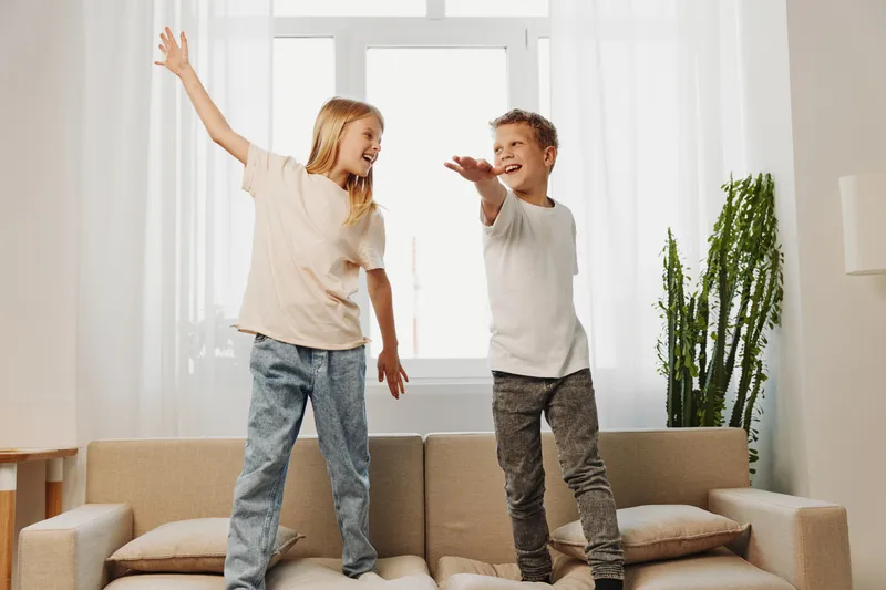 Happy children stand on sofa in bright living room, smiling and playing together. Casual clothes and natural light create joyful and lively home atmosphere with family fun concept.