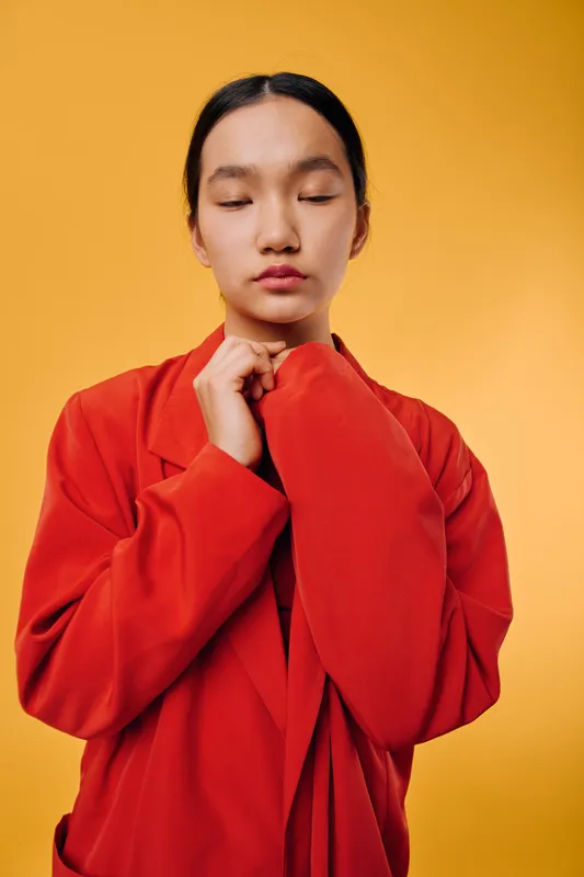 Young woman with closed eyes wearing red coat standing in front of yellow background calm expression portrait minimal studio fashion style with natural skin and dark hair tied back.